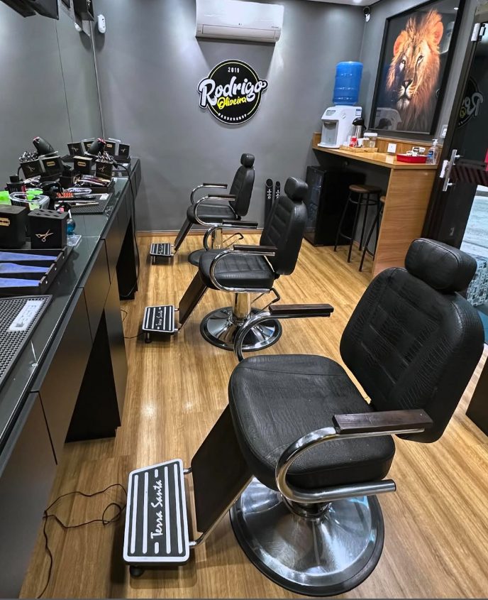 Salão da barbearia com três cadeiras pretas, piso de madeira e quadro de leão na parede